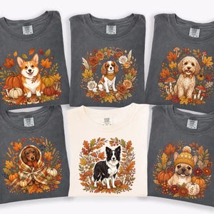 May include: Five charcoal gray and one cream-colored t-shirt featuring autumn-themed graphic prints of various dog breeds including a Corgi, Cavalier King Charles Spaniel, Doodle, Dachshund, Border Collie, and French Bulldog surrounded by fall foliage.