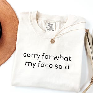 May include: Cream-colored t-shirt with the text "sorry for what my face said" in black. A gold necklace with a pendant and a brown hat are also visible. The shirt has a Comfort Colors tag.