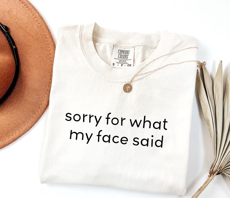 May include: Cream-colored t-shirt with the text "sorry for what my face said" in black. A gold necklace with a pendant and a brown hat are also visible. The shirt has a Comfort Colors tag.