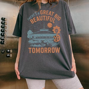 May include: Dark gray t-shirt with the text "THERE'S A GREAT BIG BEAUTIFUL TOMORROW" in orange and white. The graphic includes a building, sun, and gears in blue and orange. The shirt is worn by a person.