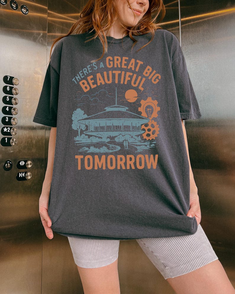 May include: Dark gray t-shirt with the text "THERE'S A GREAT BIG BEAUTIFUL TOMORROW" in orange and white. The graphic includes a building, sun, and gears in blue and orange. The shirt is worn by a person.
