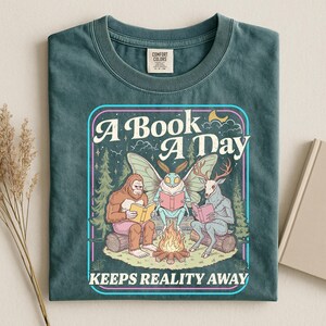 May include: A dark teal t-shirt featuring a graphic print of a Bigfoot, a moth-like creature, and a deer-headed figure sitting around a campfire reading books. The text reads A BOOK A DAY KEEPS REALITY AWAY. The shirt has a crew neck and short sleeves.