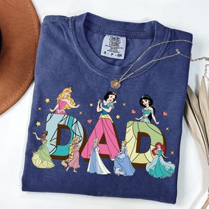 May include: A navy blue t-shirt featuring the word "DAD" with Disney princess illustrations. The letters are in a brown and blue design, with each letter featuring a different princess. The shirt has a Comfort Colors tag.