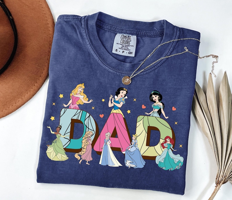 May include: A navy blue t-shirt featuring the word "DAD" with Disney princess illustrations. The letters are in a brown and blue design, with each letter featuring a different princess. The shirt has a Comfort Colors tag.