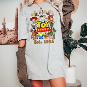 May include: A light gray t-shirt featuring a colorful graphic of characters from the animated film "Toy Story." The design includes Woody, Buzz Lightyear, and other characters, with the text "TOY STORY" and "Est. 1995."
