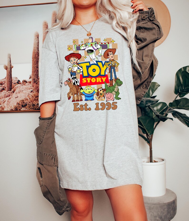 May include: A light gray t-shirt featuring a colorful graphic of characters from the animated film "Toy Story." The design includes Woody, Buzz Lightyear, and other characters, with the text "TOY STORY" and "Est. 1995."
