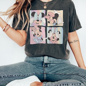 Op de afbeelding: Donkergrijs T-shirt met een ontwerp met vier panelen met Minnie Mouse-illustraties. Elk paneel heeft een pastelkleurige regenboogachtergrond. Het shirt wordt gedragen met een blauwe jeans en witte sneakers.