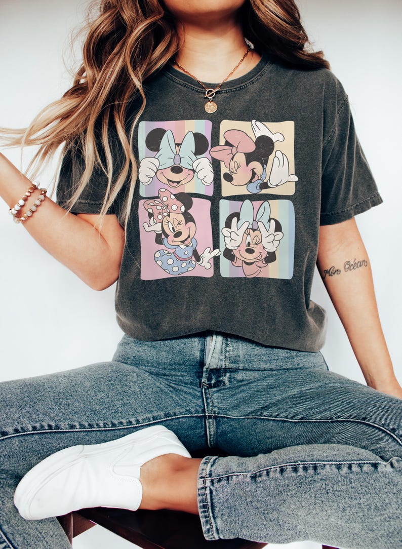 Op de afbeelding: Donkergrijs T-shirt met een ontwerp met vier panelen met Minnie Mouse-illustraties. Elk paneel heeft een pastelkleurige regenboogachtergrond. Het shirt wordt gedragen met een blauwe jeans en witte sneakers.