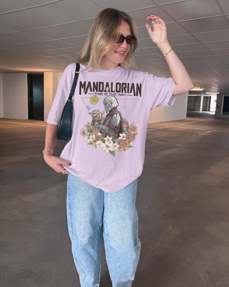 May include: A light purple short-sleeve t-shirt featuring a graphic print of The Mandalorian and Grogu surrounded by colourful flowers. The text reads MANDALORIAN THIS IS THE WAY. The shirt is paired with light blue denim jeans and a black shoulder bag.