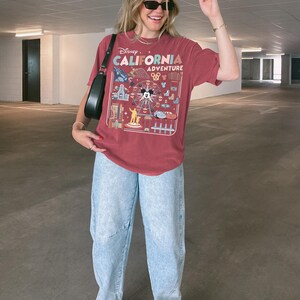 Op de afbeelding: Een bordeauxrood T-shirt met een afbeelding van het Disney California Adventure themapark. Het shirt heeft een reuzenrad, Mickey Mouse en andere attracties. De persoon draagt een lichtblauwe jeans en witte sneakers.