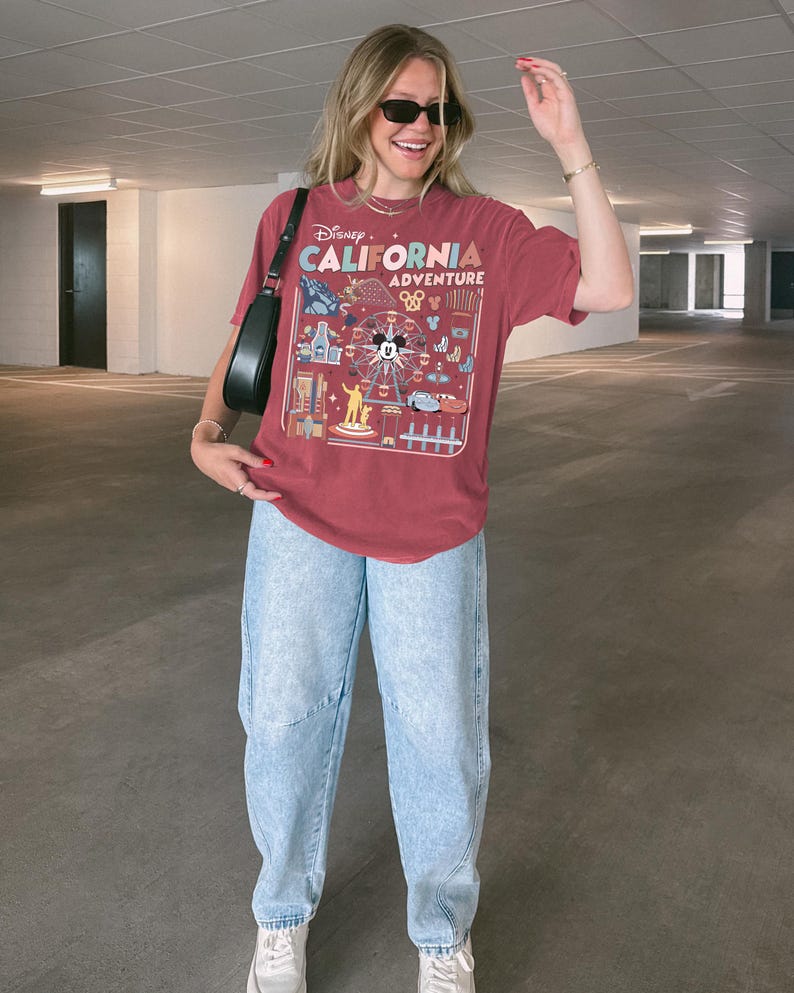 Op de afbeelding: Een bordeauxrood T-shirt met een afbeelding van het Disney California Adventure themapark. Het shirt heeft een reuzenrad, Mickey Mouse en andere attracties. De persoon draagt een lichtblauwe jeans en witte sneakers.