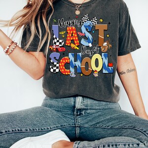Peut inclure: T-shirt gris fonc&eacute; avec l'inscription "Happy Last Day of School" dans des lettres color&eacute;es et ludiques. Le motif comprend des voitures de course, un drapeau &agrave; damier et d'autres graphiques sur le th&egrave;me de l'&eacute;cole. La personne porte un jean bleu et des baskets blanches.