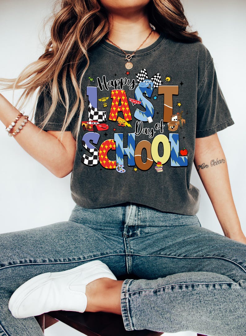 Peut inclure: T-shirt gris fonc&eacute; avec l'inscription "Happy Last Day of School" dans des lettres color&eacute;es et ludiques. Le motif comprend des voitures de course, un drapeau &agrave; damier et d'autres graphiques sur le th&egrave;me de l'&eacute;cole. La personne porte un jean bleu et des baskets blanches.