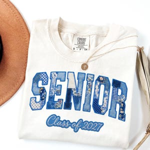 以下が含まれることがあります： クリーム色のComfort Colors Tシャツ。青色のパッチワーク文字で「SENIOR」、筆記体で「Class of 2027」と書かれています。 金色のネックレスと茶色の帽子も写っています。