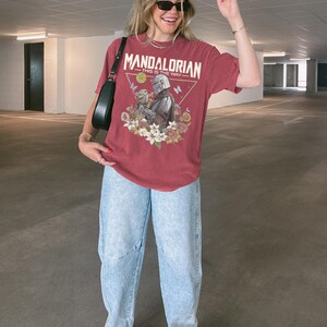 May include: A faded red short-sleeve t-shirt featuring a graphic print of The Mandalorian and Grogu, surrounded by flowers and butterflies. The text reads "MANDALORIAN" and "THIS IS THE WAY" above the characters. The shirt is paired with light blue jeans.