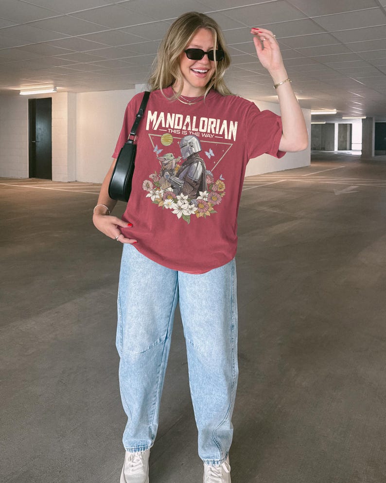 May include: A faded red short-sleeve t-shirt featuring a graphic print of The Mandalorian and Grogu, surrounded by flowers and butterflies. The text reads "MANDALORIAN" and "THIS IS THE WAY" above the characters. The shirt is paired with light blue jeans.