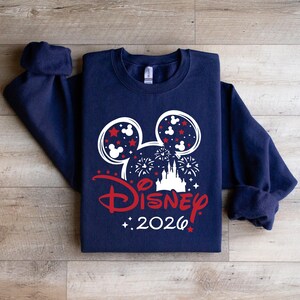 May include: Navy blue sweatshirt featuring a white and red Disney design. The design includes Mickey Mouse ears, fireworks, a castle, and the word "Disney" in red, with "2026" below. The sweatshirt is folded on a wooden surface.