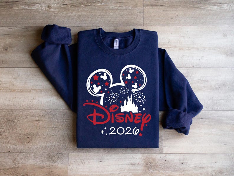 May include: Navy blue sweatshirt featuring a white and red Disney design. The design includes Mickey Mouse ears, fireworks, a castle, and the word "Disney" in red, with "2026" below. The sweatshirt is folded on a wooden surface.