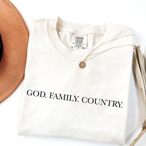 May include: Cream-colored t-shirt with the words "GOD. FAMILY. COUNTRY." printed in black. A gold necklace with a pendant rests on the shirt. A brown hat and dried plant are in the background.