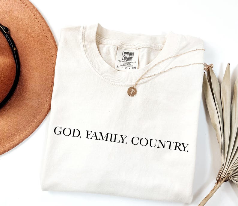 May include: Cream-colored t-shirt with the words "GOD. FAMILY. COUNTRY." printed in black. A gold necklace with a pendant rests on the shirt. A brown hat and dried plant are in the background.