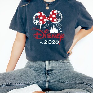 May include: Dark blue t-shirt featuring a Minnie Mouse bow with red polka dots and white accents. The word "Disney" is in red script, with "2026" below. Fireworks and a castle silhouette complete the design.