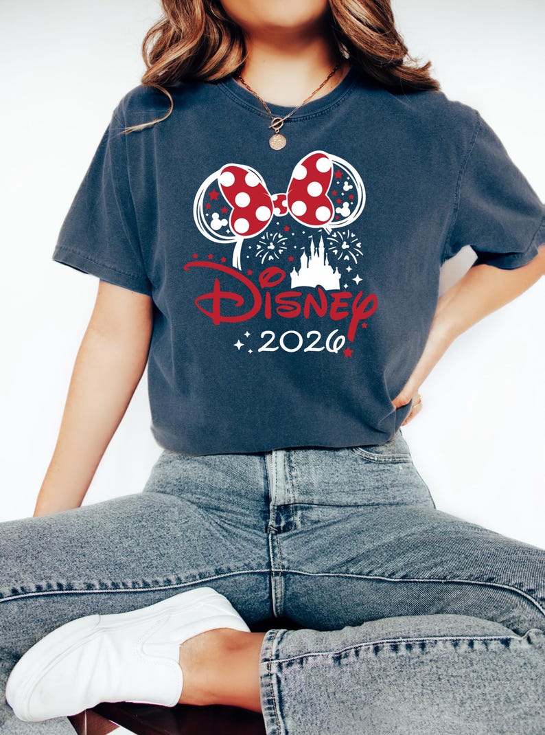 May include: Dark blue t-shirt featuring a Minnie Mouse bow with red polka dots and white accents. The word "Disney" is in red script, with "2026" below. Fireworks and a castle silhouette complete the design.