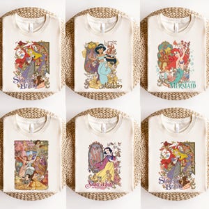 May include: A collection of six cream-colored t-shirts, each featuring a different graphic print of a classic animated character. The designs include Sleeping Beauty, Aladdin, The Little Mermaid, a group of princesses, and Snow White with floral motifs.