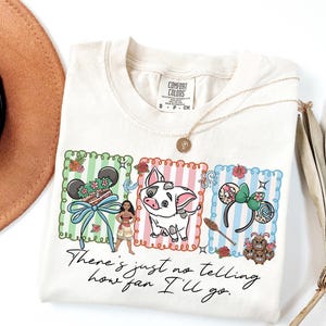 May include: A cream-colored t-shirt featuring a graphic with three vertical striped panels in green, pink, and blue. The design includes a character, a pig, mouse ears, and the text "There's just no telling how far I'll go." The shirt has a Comfort Colors label.
