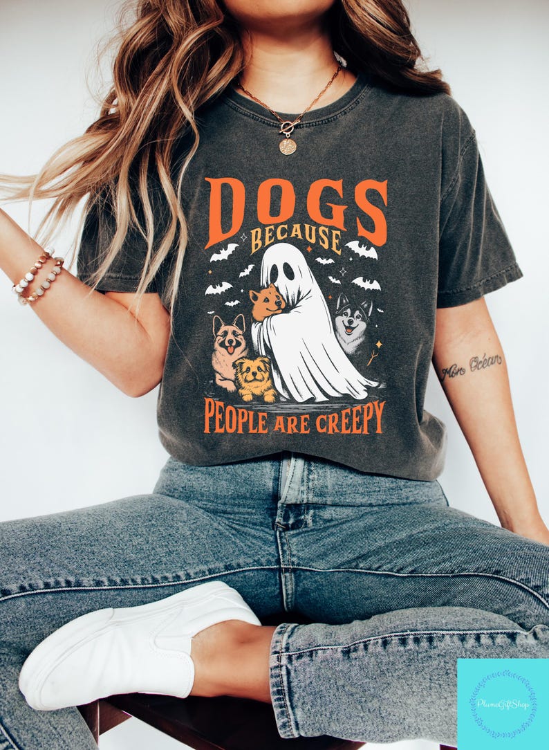 Może przedstawiać: Ciemnoszara koszulka z napisem "DOGS BECAUSE PEOPLE ARE CREEPY" w kolorze pomarańczowym i białym. Grafika przedstawia ducha trzymającego psy, z nietoperzami w tle. Koszulka ma swobodny, vintage'owy styl.