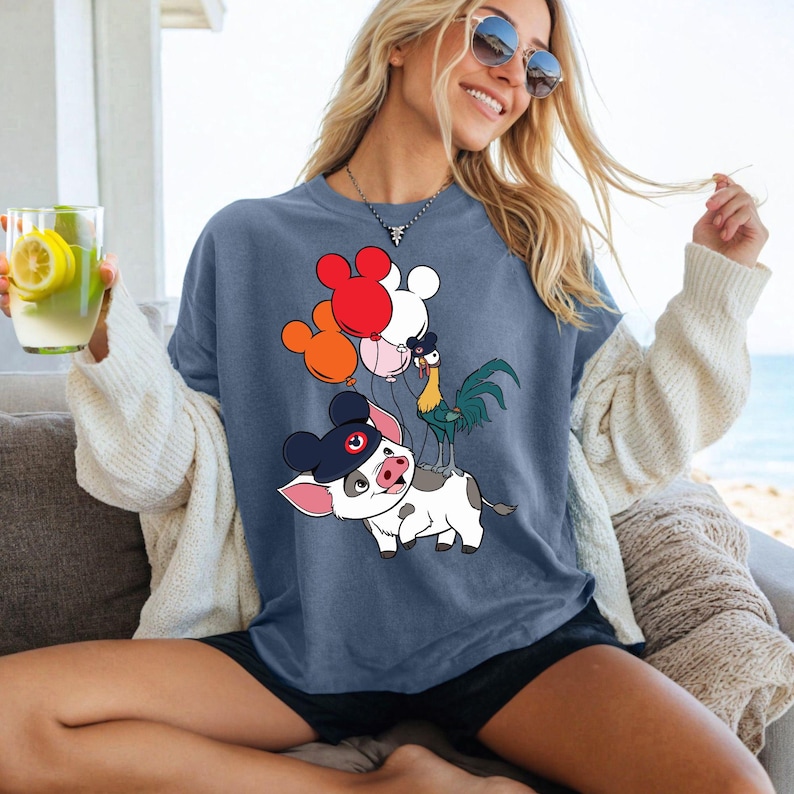 Op de afbeelding: Blauw t-shirt met een cartoonvarken met een hoed, een haan en Mickey Mouse-vormige ballonnen in rood, oranje en wit. Het varken heeft zwart-witte vlekken. De haan is groen en geel. De ballonnen worden door de haan vastgehouden.