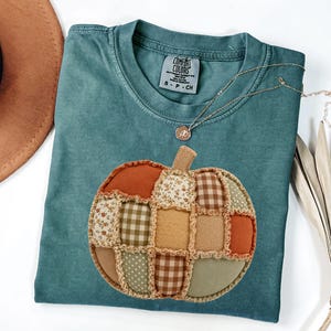May include: A teal short-sleeved t-shirt featuring a large, central pumpkin appliqué made of various fabric patches in shades of orange, brown, tan, and sage green. The shirt has a Comfort Colors label at the neck and a small rose pendant necklace.