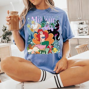 May include: A light blue t-shirt featuring a colorful graphic from "The Little Mermaid." The design includes Ariel, King Triton, Ursula, and other characters. The shirt is worn by a person sitting on a white surface.