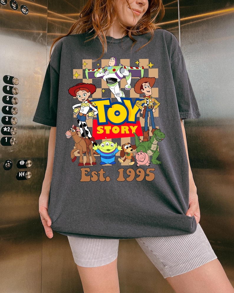 May include: Dark gray t-shirt featuring a colorful graphic of Toy Story characters, including Woody, Buzz Lightyear, and Jessie. The words "Toy Story" are in a yellow and red font, with "Est. 1995" below. The shirt has short sleeves and a relaxed fit.