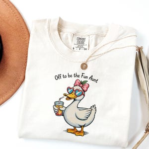 May include: A cream-coloured t-shirt featuring a graphic of a duck wearing heart-shaped sunglasses and a pink bow, holding a glass of orange juice. The text above the duck reads, Off to be the Fun Aunt. The shirt has a Comfort Colors brand label.