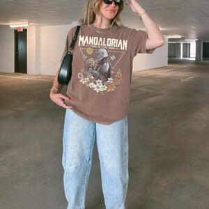 May include: A brown short-sleeve t-shirt featuring a graphic print of The Mandalorian and Grogu surrounded by colourful flowers and butterflies. The text "MANDALORIAN" and "THIS IS THE WAY" is printed above the characters. Light blue acid-wash jeans.