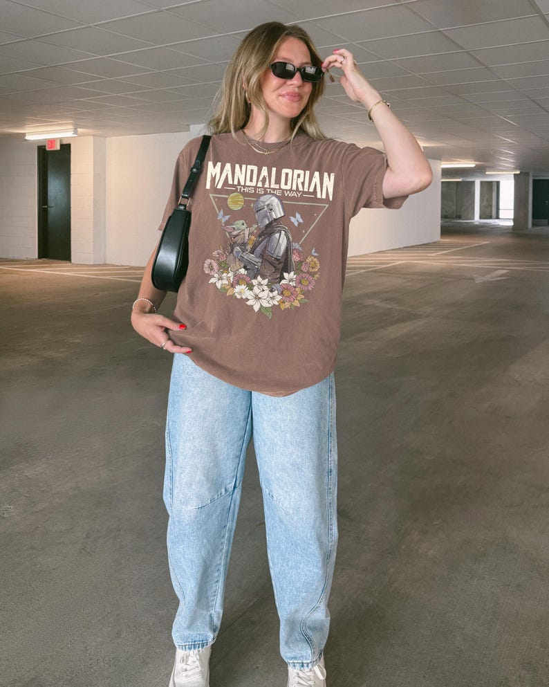 May include: A brown short-sleeve t-shirt featuring a graphic print of The Mandalorian and Grogu surrounded by colourful flowers and butterflies. The text "MANDALORIAN" and "THIS IS THE WAY" is printed above the characters. Light blue acid-wash jeans.