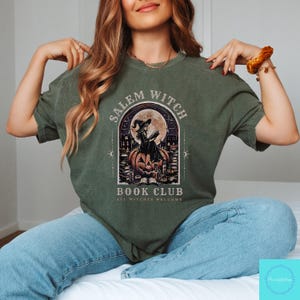May include: Olive green t-shirt featuring a graphic for the Salem Witch Book Club. The design includes a witch reading on a pumpkin, a full moon, and the text "ALL WITCHES WELCOME". The shirt is a crew neck style.