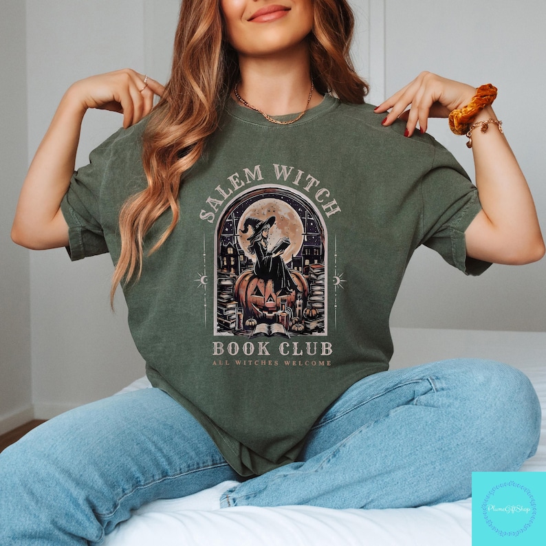 May include: Olive green t-shirt featuring a graphic for the Salem Witch Book Club. The design includes a witch reading on a pumpkin, a full moon, and the text "ALL WITCHES WELCOME". The shirt is a crew neck style.