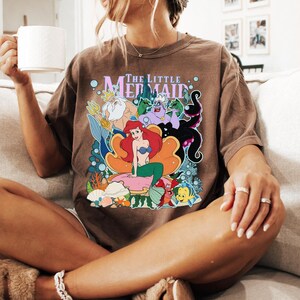May include: Brown t-shirt featuring a colorful graphic of Ariel and other characters from "The Little Mermaid". The shirt has the text "The Little Mermaid" above the graphic. The person is sitting on a beige couch.