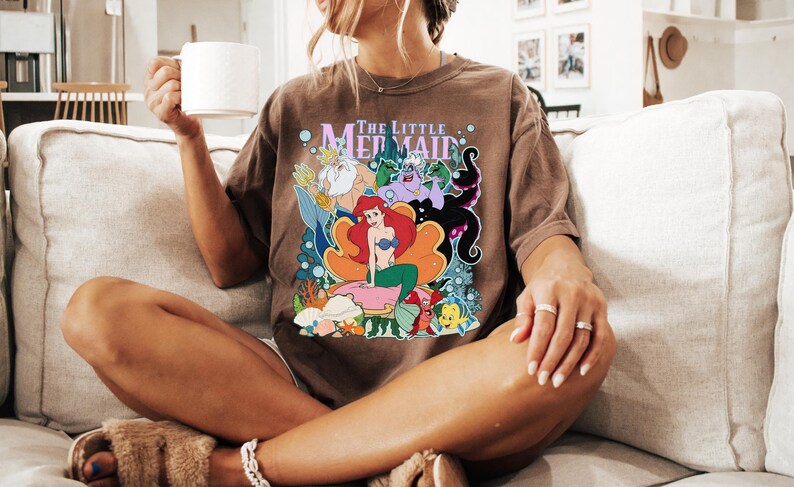 May include: Brown t-shirt featuring a colorful graphic of Ariel and other characters from "The Little Mermaid". The shirt has the text "The Little Mermaid" above the graphic. The person is sitting on a beige couch.