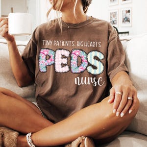 May include: A brown t-shirt with the text "Tiny Patients, Big Hearts" above the word "PEDS" in a floral and striped design, and the word "nurse" below. The shirt is worn by a person sitting on a couch.