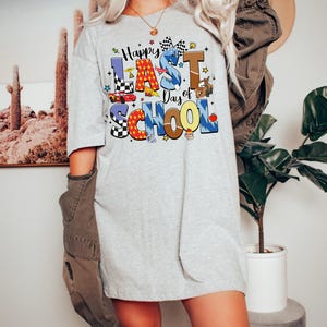 Peut inclure: Un t-shirt gris clair avec l'inscription "Happy Last Day of School" en lettres color&eacute;es et ludiques. Le motif comprend des voitures de course, des &eacute;toiles et une pomme. Le t-shirt est de coupe ample et d&eacute;contract&eacute;e.