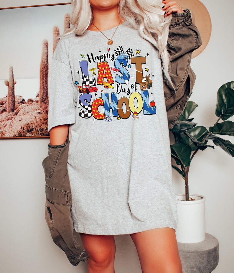 Peut inclure: Un t-shirt gris clair avec l'inscription "Happy Last Day of School" en lettres color&eacute;es et ludiques. Le motif comprend des voitures de course, des &eacute;toiles et une pomme. Le t-shirt est de coupe ample et d&eacute;contract&eacute;e.