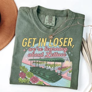 Może przedstawiać: Oliwkowy t-shirt z nadrukiem graficznym przedstawiającym przejażdżkę łodzią przez szklarnię z sałatą. Tekst brzmi "GET IN LOSER, we're learning about Lettuce". Koszulka ma okrągły dekolt i metkę marki Comfort Colors przy kołnierzu.