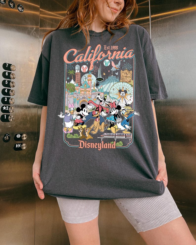 Op de afbeelding: Donkergrijs T-shirt met een Disneyland-afbeelding. De afbeelding bevat de tekst "California Est. 1955" en afbeeldingen van Mickey Mouse, Minnie Mouse, Donald Duck en andere Disney-personages. Het T-shirt heeft een losse pasvorm.