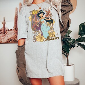 May include: A light gray t-shirt featuring a graphic print of Princess Jasmine from Aladdin, surrounded by yellow roses, small birds, and the word Aladdin in a cursive script. The shirt has short sleeves and a crew neckline, worn with a dark jacket.