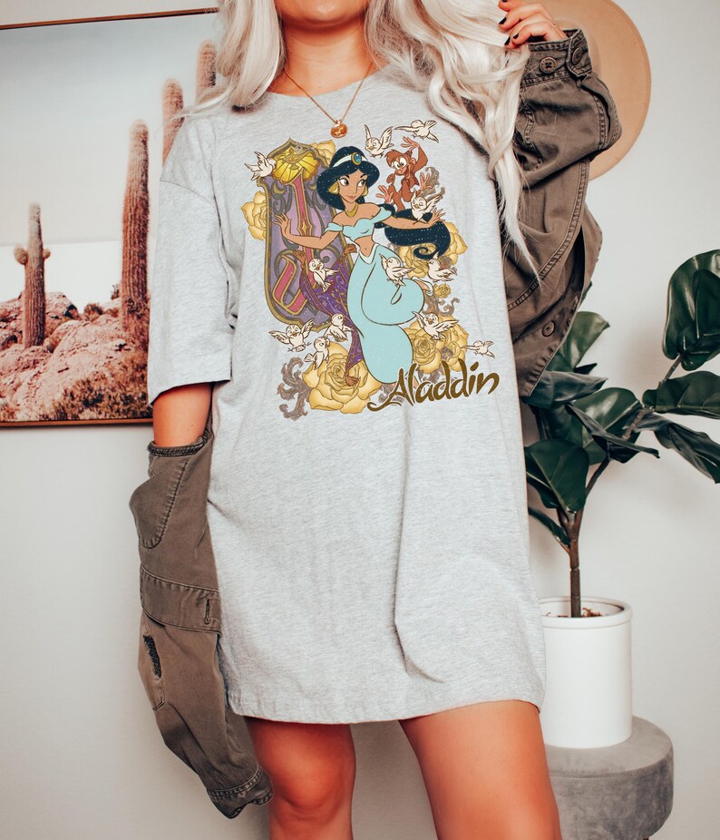 May include: A light gray t-shirt featuring a graphic print of Princess Jasmine from Aladdin, surrounded by yellow roses, small birds, and the word Aladdin in a cursive script. The shirt has short sleeves and a crew neckline, worn with a dark jacket.