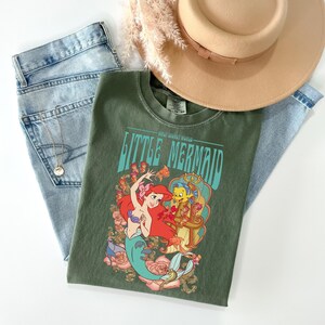May include: An olive green t-shirt featuring the text "Little Mermaid" with a graphic of Ariel and Flounder. The shirt is paired with light wash denim jeans, a gold necklace, and a tan hat with a brown band. Dried flowers are also present.