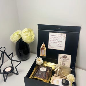 May include: A black gift box with a gold interior, containing dates, honey, a small book, and a digital counter. The box is decorated with white roses and a sticker with text. A black vase with white flowers is to the left.