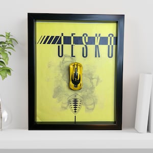 May include: A framed artwork with a yellow background and the word "JESKO" in black. A yellow toy car is mounted in front of the background, with the Koenigsegg logo below. The frame is black.